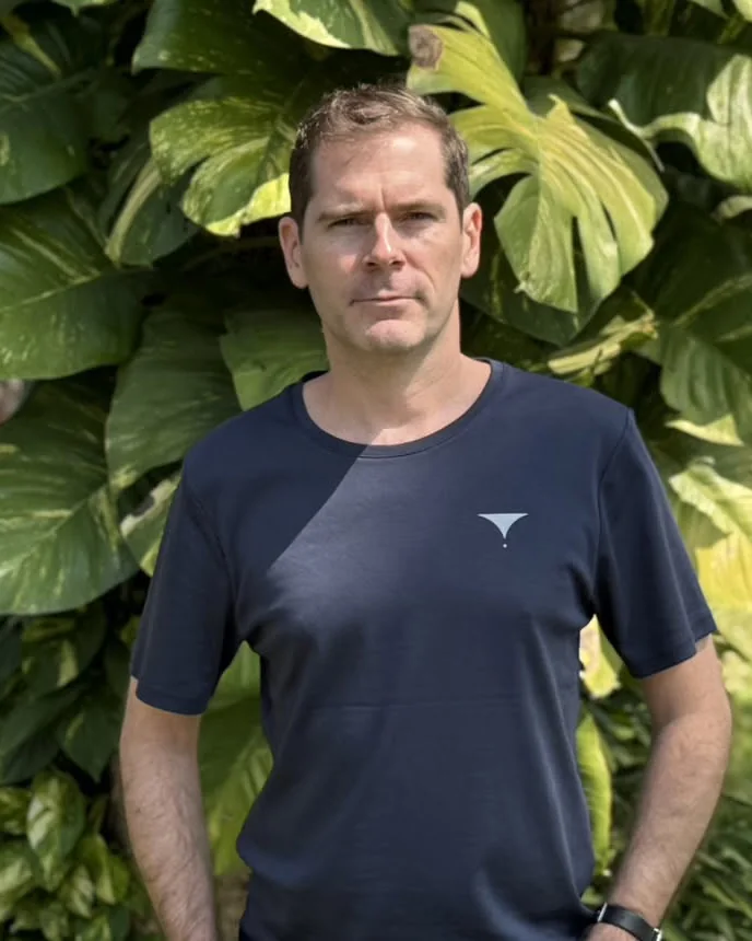 Sébastien in a navy Essential Studio T-shirt, standing in front of large monstera leaves in a sunny garden.