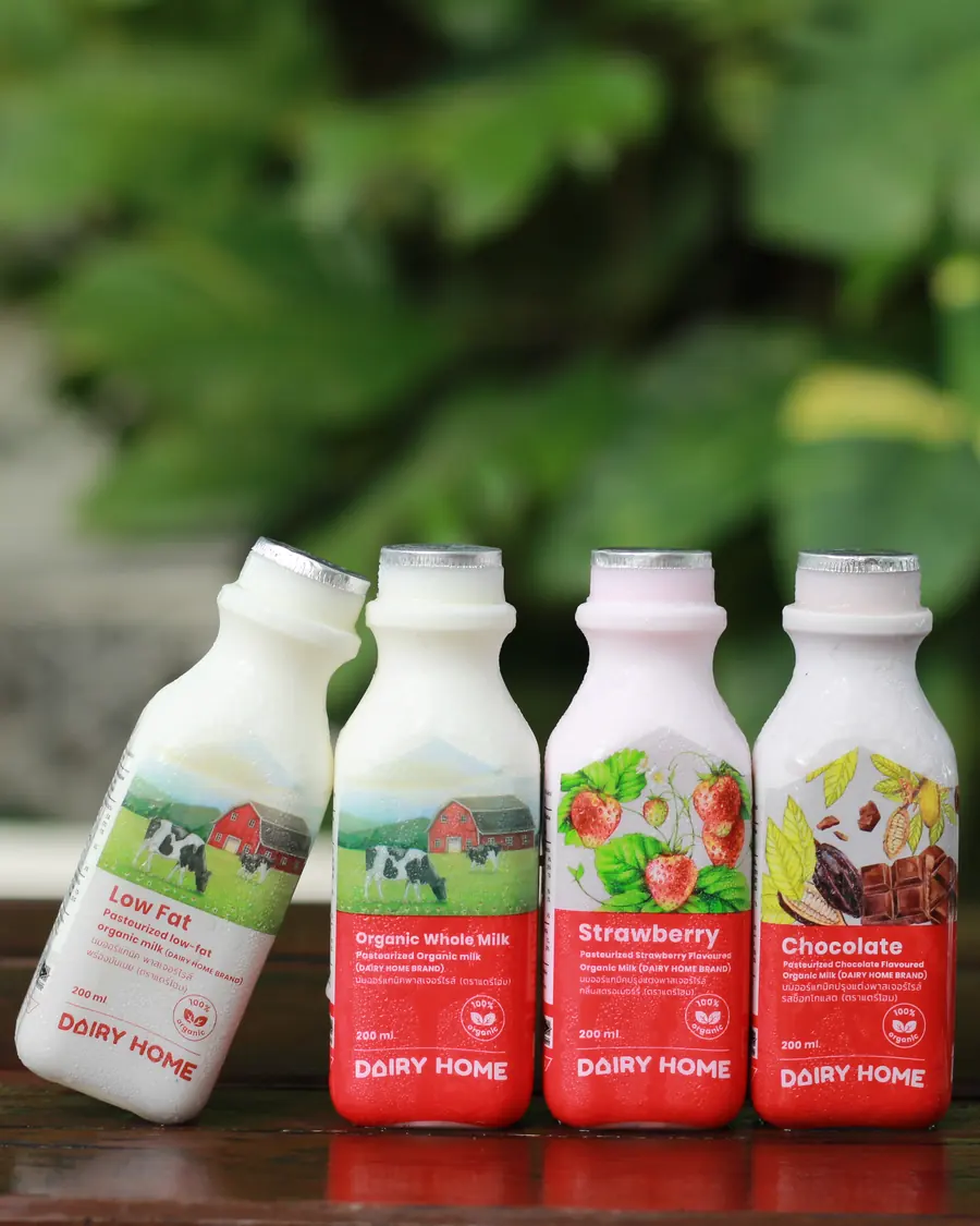 Four Dairy Home milk bottles with farm and fruit labels on a wooden table, greenery behind.