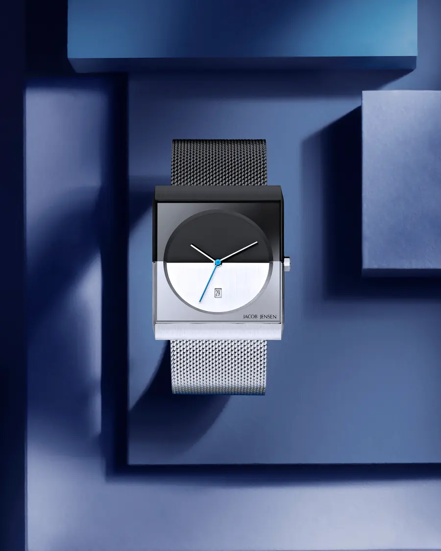A square Jacob Jensen watch with a black and white split dial and a steel mesh strap, on a deep blue set.