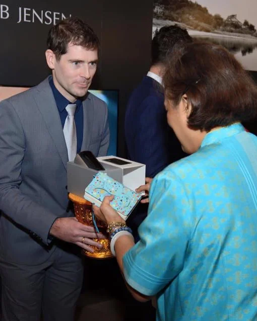 Sébastien, in a grey suit, presenting a product in its box to Her Royal Highness Princess Maha Chakri Sirindhorn at the Jacob Jensen Design studio.