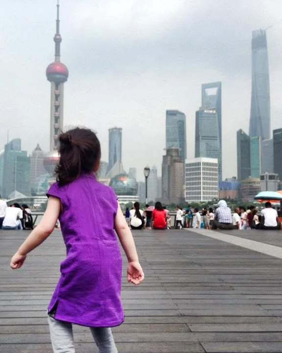 A little girl in a purple dress, seen from behind, running along the Bund boardwalk towards the Pudong skyline and the Oriental Pearl Tower.