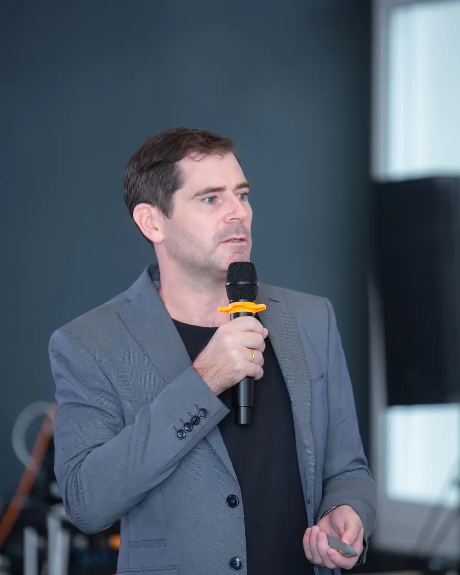 Sébastien speaking into a handheld microphone, in a grey blazer.